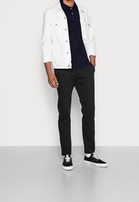 Vit denimjacka, marinblå polo, svarta byxor och svarta sneakers med vita detaljer och snören, uppvisade mot en ljus bakgrund.