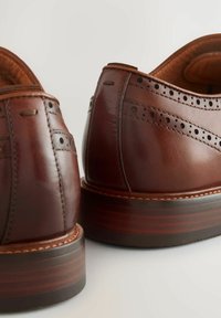 Pantofi din piele maro cu detalii brogue și un finisaj neted, având un toc din lemn stratificat și cusături în contrast.