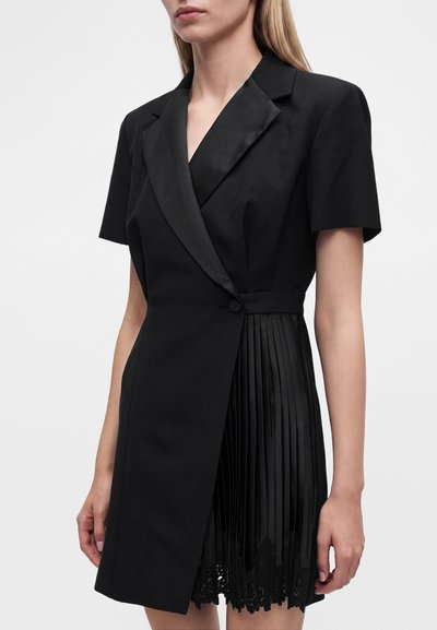 Femme portant une robe blazer noire à manches courtes avec un col en satin, une fermeture à un bouton et une jupe plissée avec un bord en dentelle.