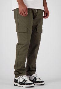 Olijfgroene cargobroeken met een taps toelopende pasvorm, meerdere zijzakken en elastische boorden, gecombineerd met zwart-witte sneakers.