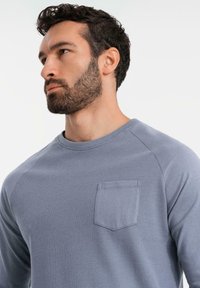 Graues Langarmhemd mit strukturiertem Körper und glatten Ärmeln, mit einer Fronttasche und einem Rundhalsausschnitt.