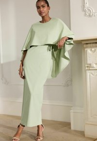 Robe vert clair avec un superposé ressemblant à une cape, silhouette ajustée, texture lisse et design long au sol. Complétée par des sandales à talons hauts beiges.