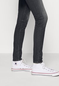 Jeans skinny neri con una texture liscia, caratterizzati da dettagli con bottoni alla caviglia. Abbinati a sneakers bianche alte con rifiniture rosse.