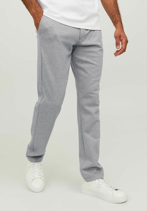 Man draagt lichtgrijze sweatpants en witte sneakers, met één hand in zijn zak, staande tegen een lichte achtergrond.