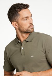 Polo di colore verde oliva con un tessuto testurizzato, tre bottoni al colletto e un piccolo logo bianco sul lato sinistro del petto. Maniche corte.