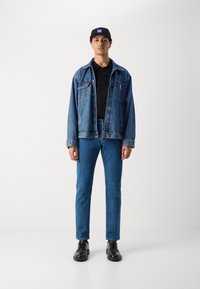 Ung mand iført en sort cap, sort polo, blå denimjakke, blå jeans og sorte lædersko, stående foran en ensfarvet hvid baggrund.
