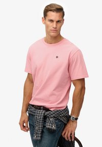 Roze katoenen T-shirt met korte mouwen en een ronde hals, voorzien van een klein zwart logo op de borst. Gecombineerd met een blauwe spijkerbroek en een geruit overhemd dat rond de taille is gestrikt.