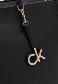Sac à main en cuir texturé noir avec des équipements en or. Il dispose d'une bandoulière amovible et d'un accent doré "ck" bien en vue.