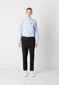 Lichtblauwe zip-up jas met hoge kraag en zwarte accenten, gecombineerd met zwarte broek en witte sneakers. De jas heeft een geborduurd logo.