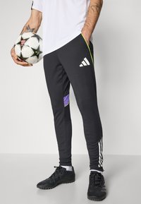 Zwarte sportbroeken met witte strepen, paarse en groene accenten, gemaakt van zacht materiaal. Bevestigd aan een voetbalschoen, gedragen met zwarte voetbalschoenen.