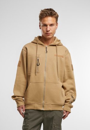 Tan zip-up hoodie med snoretræk, frontlomme med lynlås, kontrasterende patch på brystet og ribbede manchetter. Stoffet ser blødt og tekstureret ud.