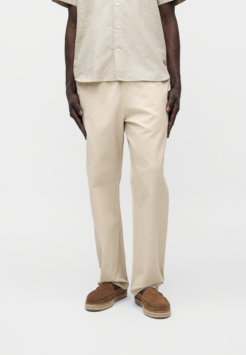 Homme portant un pantalon beige décontracté, une chemise à manches courtes beige clair à boutons, et des chaussures marron à enfiler avec des semelles tissées.