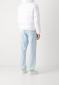 Osoba ubrana w białą kurtkę puchową, jasnoniebieskie dżinsy o prostym kroju i białe sneakersy, stojąca na jednolitym jasnym tle.