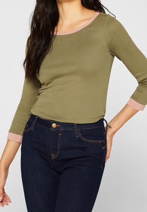 Nainen, joka pukeutunut oliivinvihreään pitkähihainen paitaan, jossa on vaaleanpunainen reunus, ja tummansinisiin farkkuihin, poseeraa käsi lanteillaan.