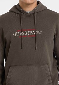 Felpa marrone con tasca frontale e cordini, con il testo ricamato in rosso e bianco "GUESS JEANS" sul petto.