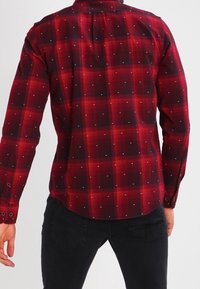 Camisa de cuadros roja y negra con mangas largas, con detalles de puntos plateados, cuello abotonado y dobladillo curvado.