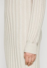Hand rustend naast het lichaam, gekleed in een crèmekleurige geribde trui met lange mouwen en manchetdetails.