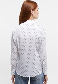 White long-sleeve shirt featuring a small blue circular pattern, with a rounded hem and buttoned cuffs. Smooth, lightweight fabric.