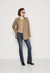 Blazer beige de doble botonadura, top ajustado color tostado, jeans azul oscuro y botas negras de tobillo. La modelo lleva gafas de sol color crema. Fondo blanco liso.
