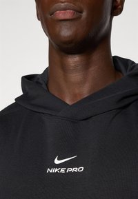 Černá mikina Nike Pro s přední kapsou, stahovací kapucí a bílými detaily loga. Hladká textura a moderní sportovní design.