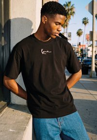 Joven con camisa negra de manga corta con logo en escritura blanca, apoyado contra una pared en una acera urbana soleada con palmeras y señales de tráfico.