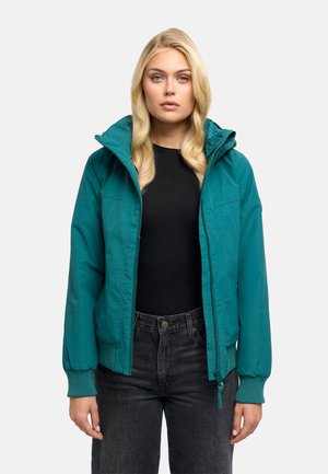 Femme blonde aux boucles lâches porte une veste zippée bleu sarcelle par-dessus un t-shirt noir et un jean foncé, debout devant un fond uni.