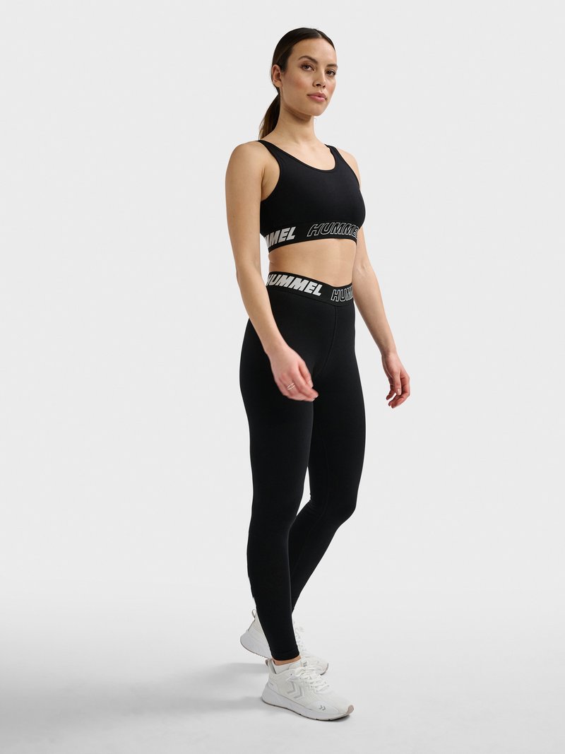Frau steht und trägt schwarzen Sport-BH und Leggings mit weißen "Hummel" Logo-Bändern und weißen Sportschuhen vor grauem Hintergrund.