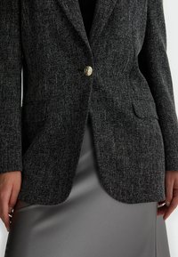 Blazer nero strutturato con un unico bottone dorato, due tasche anteriori e una vestibilità su misura, abbinato a una gonna in satin grigio liscio.