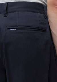 Gros plan sur un pantalon bleu marine foncé avec une poche passepoilée arrière comportant une petite étiquette de la marque Barbour sur la hanche droite.