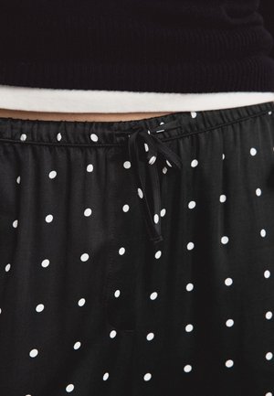 Mustat housut valkoisilla pilkkuilla, joissa on solmittava nyörivyötärö, yhdistettynä mustaan paitaan, jonka alla näkyy valkoisen aluspaidan helma.