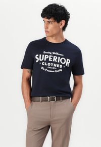Navyblauwe katoenen t-shirt met witte grafische tekst "SUPERIOR CLOTHES." Gecombineerd met lichtbruine broek en een bijpassende riem.