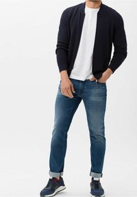 Chaqueta bomber de punto azul marino sobre una camiseta blanca, combinada con jeans ajustados azul doblados y zapatillas oscuras con acentos rojos y blancos.