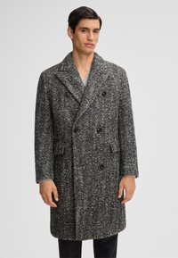 Man in een lange, dubbelzijdige zwart-witte gestructureerde wol coat met brede revers en klepzakken, staand tegen een effen achtergrond.