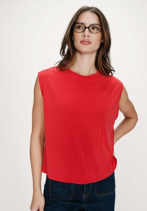 Femme portant des lunettes écaille de tortue, un haut rouge sans manches et un jean bleu foncé, debout devant un fond clair uni.