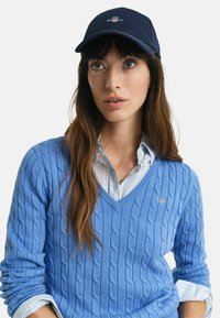 Blue cable knit sweater with a V-neck, layered over a light striped collared shirt. A navy cap with logo completes the look.