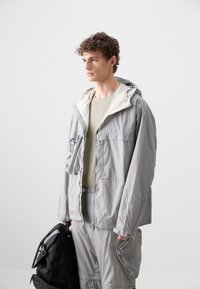 Light gray outdoor jacket with a hood, zippered front, and textured fabric. Paired with a light green shirt, holding a black backpack.