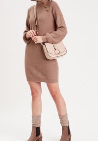 Strik beige kjole med høj krave og guld knapdetaljer, parret med en beige crossbody taske med cirkulære hardware detaljer.