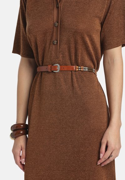 Nő, aki barna, rövid ujjú, gombos ruhát visel, vékony, világosbarna övvel és két tömzsi barna karkötővel a bal csuklóján.