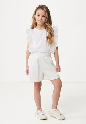 Haut blanc à manches courtes avec des manches volantes, associé à un short blanc brodé. Le modèle porte des baskets blanches avec des accents dorés.
