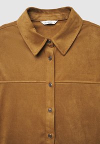 Chemise marron en daim avec un col pointu, une fermeture à boutons sur le devant et un détail de couture horizontale à travers la poitrine.