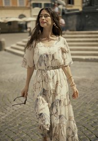 Lágy, krémszínű ruha lómintával, rövid ujjakkal, húzózsinóros derékkal és fodros szegéllyel. A modell napszemüveget tart, kövekkel kirakott úton áll.