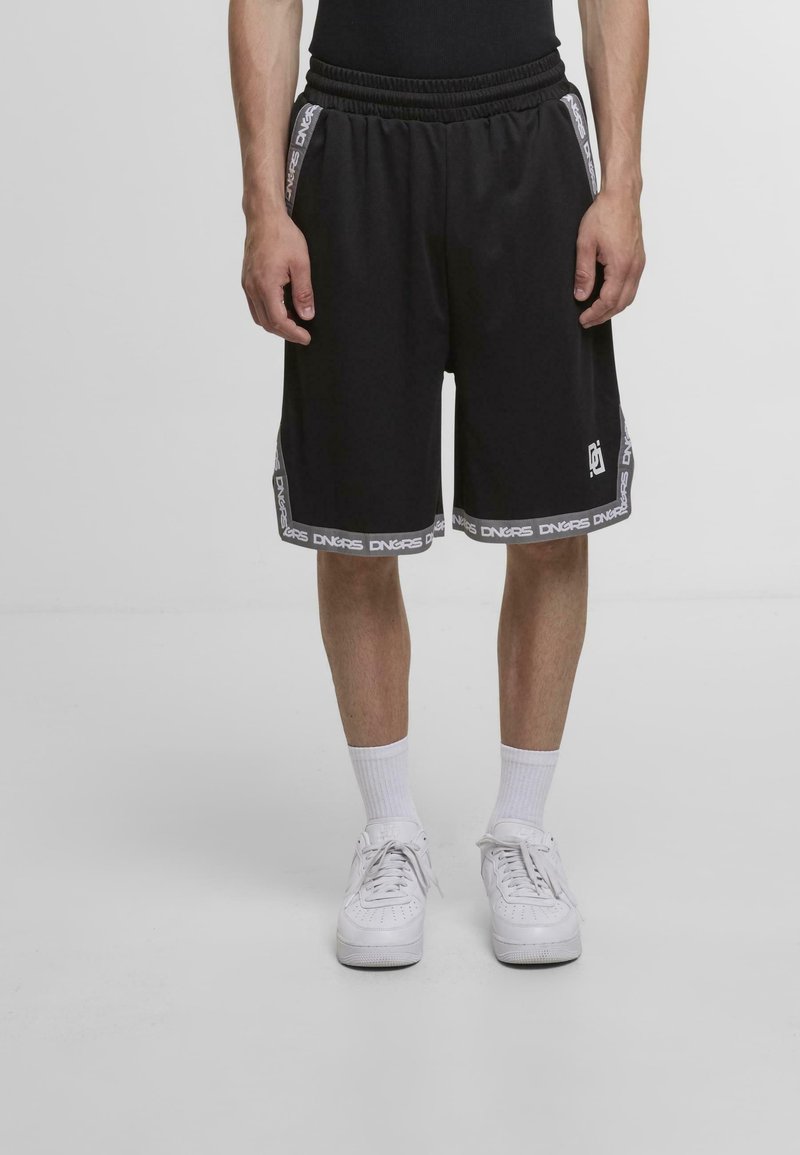 Homme portant des shorts noirs arrivant aux genoux avec des logos et des textes blancs, des chaussettes blanches et des baskets blanches, se tenant sur un sol uni.