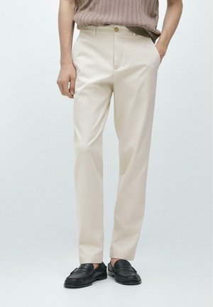 Uomo che indossa pantaloni beige a gamba dritta, un maglione marrone e mocassini neri, in piedi con una mano in tasca.