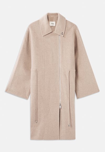 Bežový kabát z vlnenej zmesi s vysokým golierom, predným zipsom, bočnými vreckami a širokými rukávmi; hladká textúra a minimalistický dizajn.