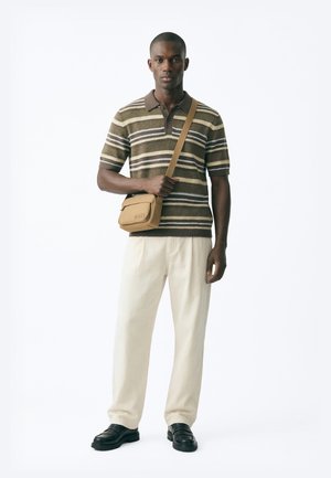 Άνδρας που φοράει καφέ ριγέ πόλο, εκρού παντελόνι, μαύρα παπούτσια και ταμπά τσάντα χιαστί, στέκεται μπροστά σε απλό λευκό φόντο.