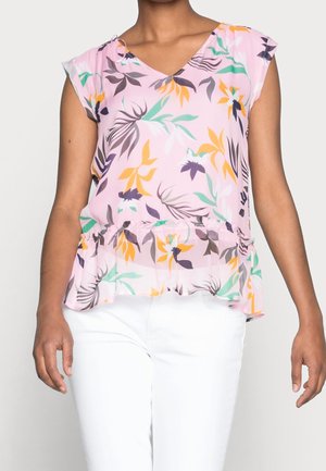 Vrouw draagt een mouwloze roze blouse met groene, gele en paarse bloemenpatronen, gecombineerd met een witte broek.
