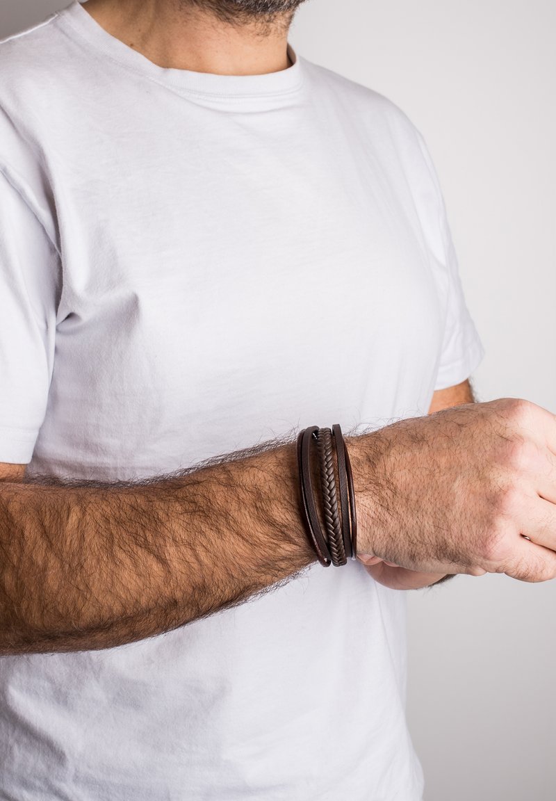 Brown leather bracelet set, featuring braided and smooth designs, worn on a wrist. The material appears durable with a textured finish.