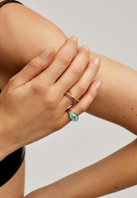 Anillos de oro y plata en una mano, con una piedra ovalada de turquesa con un diseño de cruz; piel suave y esmalte de uñas natural visibles.