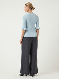 Femme aux cheveux blonds attachés en chignon, portant un pull côtelé bleu clair avec des manches à volants et un pantalon large gris foncé, debout de dos.