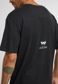 T-shirt en coton noir avec col rond, manches courtes et un petit logo blanc imprimé au dos, présentant une texture douce.